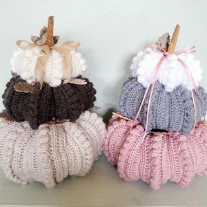 May include: Two decorative, knitted pumpkins in autumnal colours. One is beige, brown, and white, with a cinnamon stick stem and a beige ribbon. The other is pink, grey, and white, with a cinnamon stick stem and a pink ribbon.