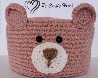 Könnte beinhalten: Ein rosa gehäkelter Korb in Bärenform mit schwarzen Augen, einer braunen Nase und einer weißen Schnauze. Der Korb ist mit einer festen Masche gehäkelt und hat eine runde Form. Der Text "By Crafty Heart" ist in der oberen rechten Ecke des Bildes sichtbar.