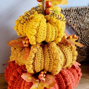 May include: A three-tiered, handmade pumpkin decoration in orange and yellow. The crocheted pumpkins are adorned with artificial leaves, berries, and a curly stem. This autumn-themed decor is ideal for adding a seasonal touch to any room.