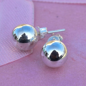 May include: A pair of shiny, silver-colored stud earrings. Each earring is a smooth, spherical shape with a post for piercing. The earrings reflect the surrounding environment, set against a soft pink background.