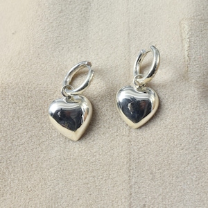 Könnte beinhalten: Ein Paar silberner herzförmiger Ohrhänger. Jeder Ohrring hat einen polierten Herzanhänger, der an einem kleinen Reifen befestigt ist. Die Ohrringe werden auf einer neutral getönten Oberfläche präsentiert.