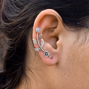 May include: Silver ear cuff featuring a floral and leaf design. The cuff curves along the ear, with a small flower detail and a pale, round stone at the top. The leaves and flower have intricate detailing.