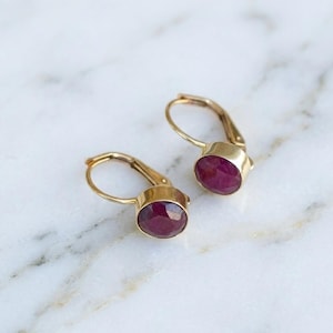 May include: A pair of gold leverback earrings, each with a deep red, oval gemstone. The earrings are displayed on a white and grey marble surface, highlighting the jewellery's design and colour.