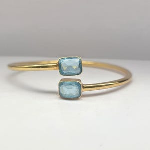 Könnte beinhalten: Ein offener Goldarmreif mit zwei rechteckigen, facettierten hellblauen Edelsteinen an den Enden. Das polierte Goldband hat eine glatte, abgerundete Oberfläche, und die beiden Steine sind einander gegenüber mit einer kleinen Lücke angeordnet.