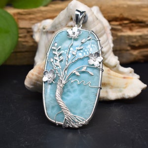 May include: A blue and silver pendant featuring a tree of life design with delicate silver flowers. The pendant is made of a blue gemstone with a silver wire wrapped around it, creating the branches and trunk of the tree. The flowers are also made of silver wire and are attached to the branches.