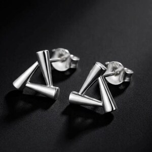 May include: A pair of silver stud earrings featuring a geometric triangular design. Each earring is constructed from three connected metallic cones forming a hollow triangle shape, complete with standard silver earring backs for secure attachment.