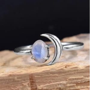 Pode incluir: Um anel de prata com um design de lua crescente segurando uma pedra preciosa oval azul clara. O anel está colocado sobre uma superfície de madeira com um fundo escuro, destacando os detalhes do anel e a cor da pedra.