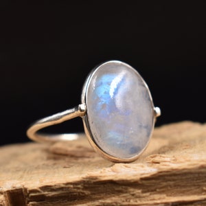 Pode incluir: Um anel de prata com uma pedra da lua oval. A pedra da lua tem uma base branca leitosa com flashes iridescentes azuis e brancos. O anel é montado em uma banda de prata simples, mostrando a beleza natural da pedra.