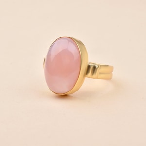 May include: A gold ring featuring a large, oval-shaped, light pink gemstone. The gemstone is set in a gold bezel, and the ring has a simple, elegant design. The ring is a piece of jewelry.