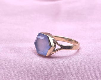 May include: A gold ring with a hexagonal, pale blue gemstone. The ring has a split-shank design. The gemstone is set in a gold bezel. The ring is displayed on a pink surface. The gemstone is a light, translucent blue.