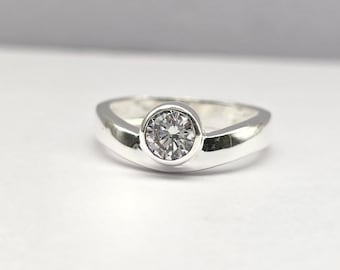 May include: A silver ring with a round, clear gemstone set in a bezel. The ring has a smooth, polished band that curves gently around the stone. The gemstone sparkles, reflecting light.