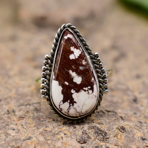 May include: A silver ring with a teardrop-shaped brown and white stone set in a silver bezel. The ring has a rope-like design around the stone.