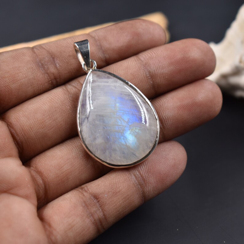 Pode incluir: Um pingente de pedra da lua em forma de l&aacute;grima com uma configura&ccedil;&atilde;o de prata. A pedra &eacute; iridescente com tons de azul e branco.