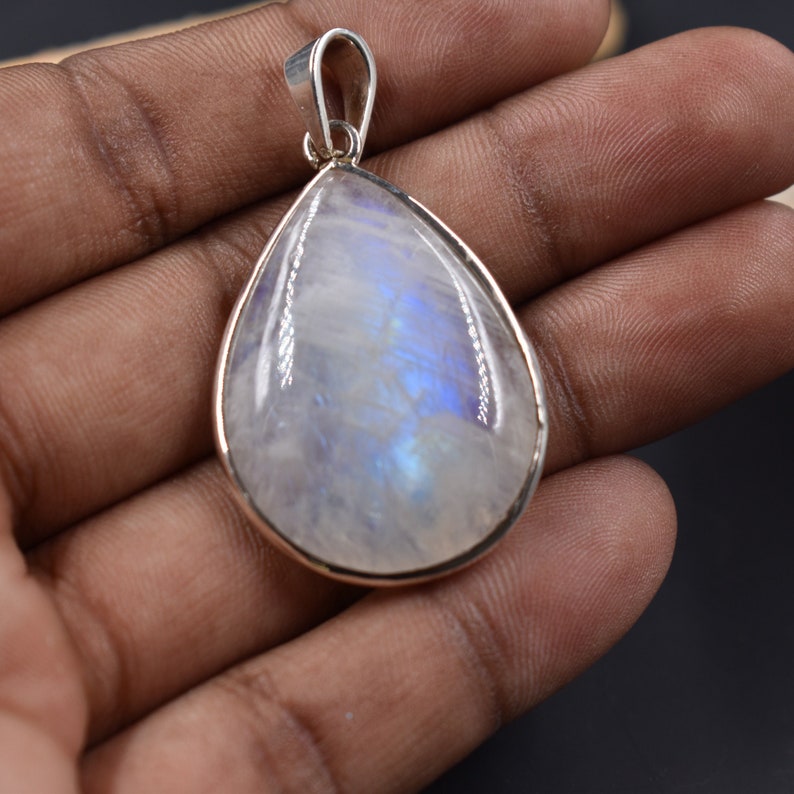 Pode incluir: Um pingente em forma de l&aacute;grima com uma configura&ccedil;&atilde;o de prata e uma grande pedra de lua iridescente. A pedra tem uma cor branca leitosa com flashes azuis e brancos.