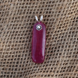 May include: A rectangular pendant with a deep red gemstone. The pendant has a silver-colored bail and a small, decorative silver accent at the top. The gemstone is smooth and polished, with a slight curve to its shape.