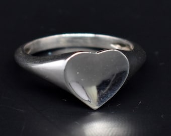 Könnte beinhalten: Ein silberner Siegelring mit einem herzförmigen Design. Der Ring ist glatt und poliert.