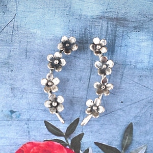 May include: A pair of silver-toned ear climbers featuring a curved design composed of four small, five-petaled flowers each. The metal has a slightly oxidized finish, highlighting the detailed petal shapes and central stamens of the floral motif.