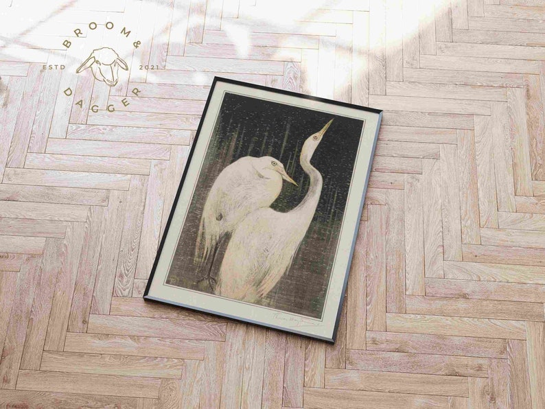 May include: A framed print of a black and white illustration of two white herons standing in a dark, watery environment. The herons are facing each other, with their necks curved and their beaks touching. The print is set against a light wood background.