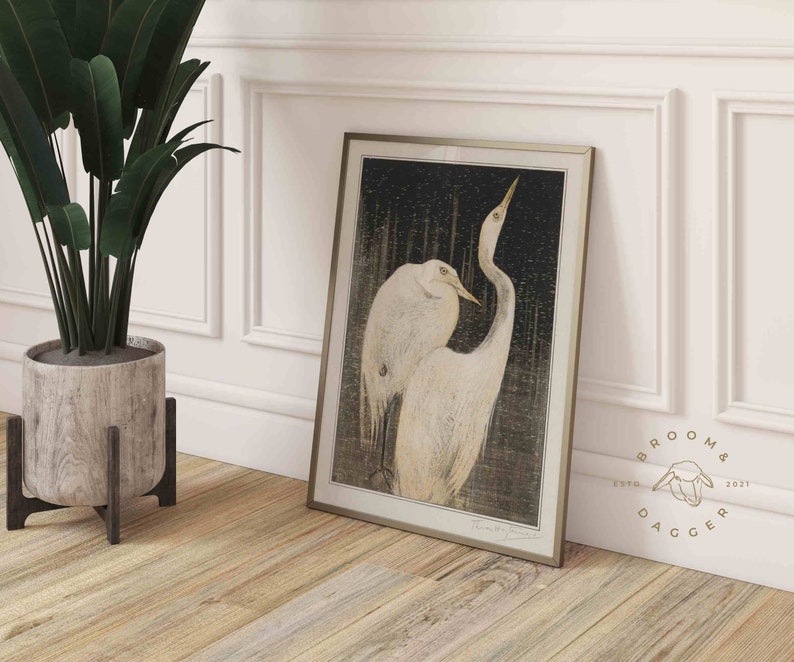 May include: A framed print of a black and white illustration of two white herons with long necks and beaks. The herons are standing in a forest setting with a dark background. The print is framed in a silver frame.