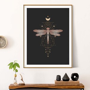 May include: A framed print with a black background and a gold geometric design featuring a dragonfly. The dragonfly is a light pink color with gold accents. The print is hanging on a white wall above a wooden console table.