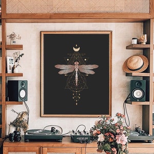 May include: A geometric dragonfly print with gold accents on a black background. The print features a crescent moon and stars. The print is framed and hanging on a wall in a room with a wooden table and shelves.