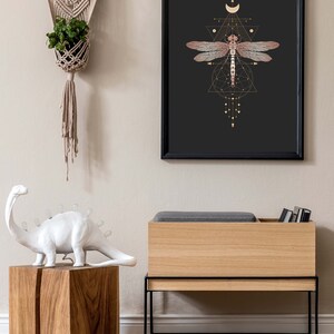 May include: A black framed print with a gold geometric design featuring a dragonfly and a crescent moon. The print is hanging on a light beige wall above a wooden storage bench with a black metal frame.