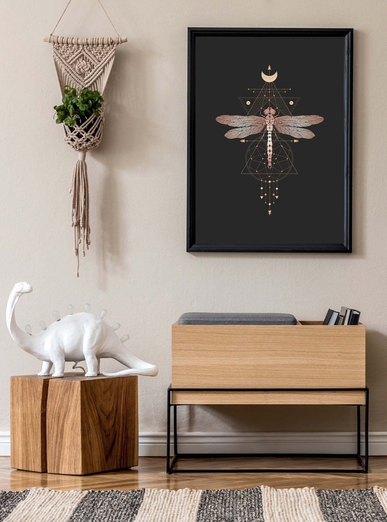 May include: A black framed print with a gold geometric design featuring a dragonfly and a crescent moon. The print is hanging on a light beige wall above a wooden storage bench with a black metal frame.