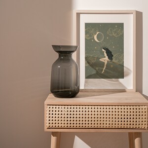 May include: A dark gray glass vase sits on a light wood table with a woven rattan drawer. Behind the vase is a framed print with a green background and a gold illustration of a woman sitting on a cloud with a crescent moon above her.