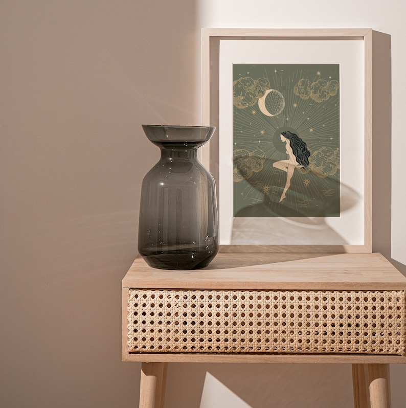 May include: A dark gray glass vase sits on a light wood table with a woven rattan drawer. Behind the vase is a framed print with a green background and a gold illustration of a woman sitting on a cloud with a crescent moon above her.