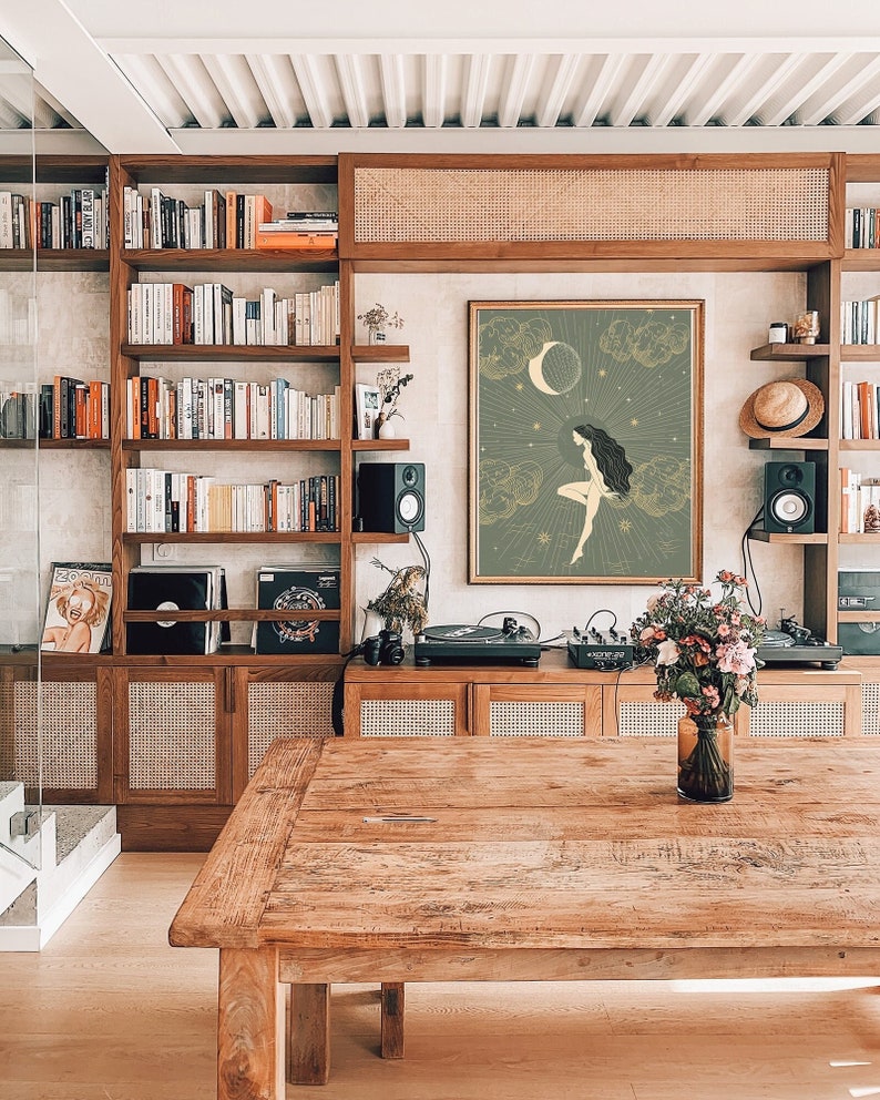 May include: A wood table with a vase of flowers on it. The table is in front of a bookshelf and a wall with a framed print of a woman standing on a cloud with a crescent moon and stars behind her.