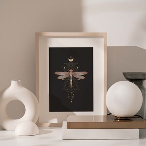 May include: A framed print of a dragonfly with geometric patterns and a crescent moon on a black background. The print is in a light wood frame and is sitting on a white surface.