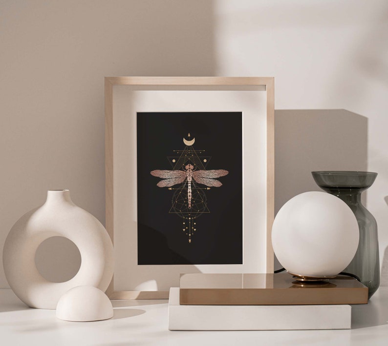 May include: A framed print of a dragonfly with geometric patterns and a crescent moon on a black background. The print is in a light wood frame and is sitting on a white surface.
