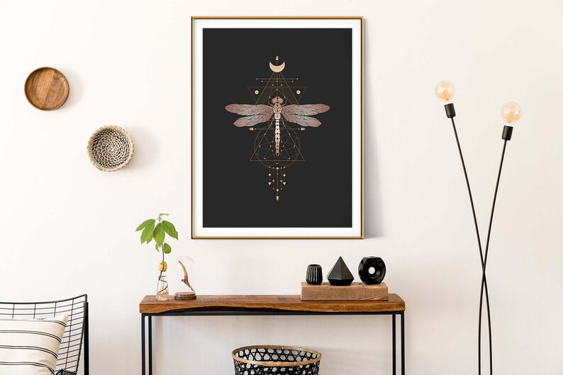 May include: A framed print with a black background and a gold geometric design featuring a dragonfly. The dragonfly is a light pink color with gold accents. The print is hanging on a white wall above a wooden console table.