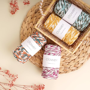 Puede incluir: Se muestra una colección de madejas de hilo coloridas, que incluyen naranja, verde, amarillo y rosa. El hilo está etiquetado como "clowex Twisted Multicolor" y está dispuesto en una cesta tejida y sobre una estera de fibra natural.