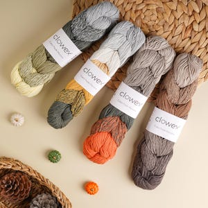 Könnte beinhalten: Vier Stränge geflochtenes Baumwollgarn in verschiedenen Farben, darunter Gelb, Grün, Orange und Grau. Jeder Strang hat ein weißes Etikett mit dem Text "clowex Cotton Puzzle Macrame". Dekorative Kürbisse und ein geflochtener Korb sind ebenfalls vorhanden.