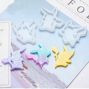 May include: A collection of Pikachu-shaped molds and finished resin figures. The molds are white, while the resin figures are in various colors, including yellow, purple, and a blue-pink gradient. The figures are on a light blue surface.