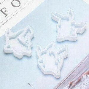 May include: Three white silicone molds in the shape of a cartoon character. The molds are arranged on a light blue surface. The molds are designed for crafting or creating resin art. The molds are detailed with the character's features.