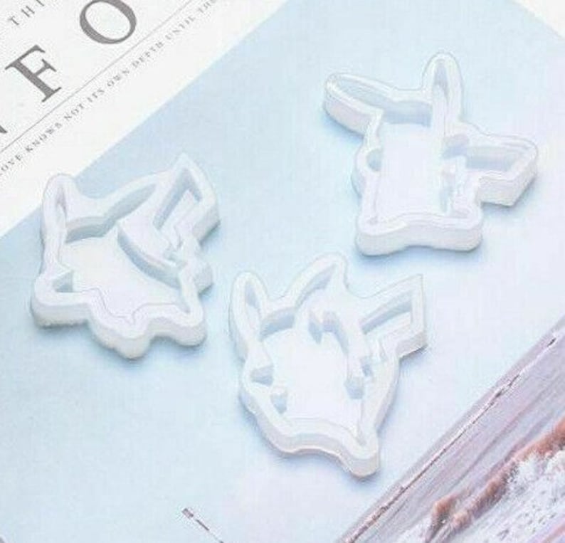 May include: Three white silicone molds in the shape of a cartoon character. The molds are arranged on a light blue surface. The molds are designed for crafting or creating resin art. The molds are detailed with the character's features.