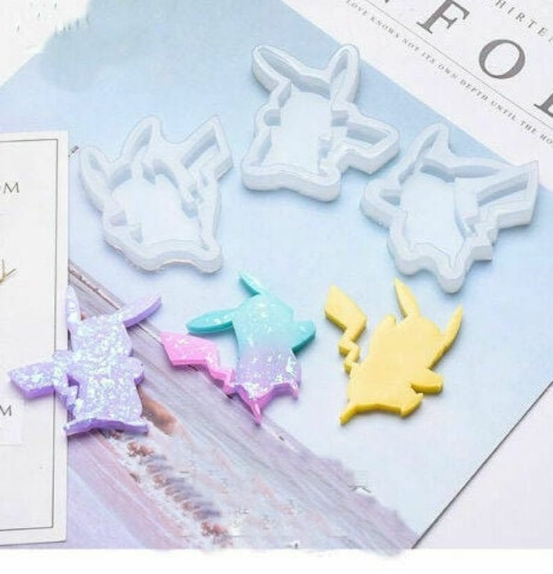 May include: A collection of Pikachu-shaped molds and finished resin figures. The molds are white, while the resin figures are in various colors, including yellow, purple, and a blue-pink gradient. The figures are on a light blue surface.