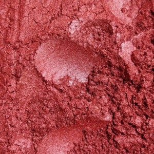 May include: A close-up shot of a textured, reddish-pink powder with lighter, pearlescent patches. The powder has a granular appearance, with visible imperfections and colour variations. The overall effect is a shimmering, metallic finish, suggesting a cosmetic product.