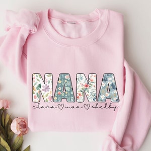 以下が含まれることがあります： 花柄の文字で「NANA」と書かれたピンク色のスウェットシャツ。文字は黒で縁取られており、「NANA」の下には黒で「clara max shelby」と書かれている。