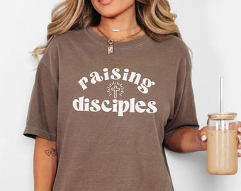 May include: A brown t-shirt with the text "raising disciples" in white lettering. The text is in a retro font and includes a cross with a sunburst behind it.