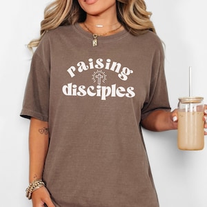 May include: A brown t-shirt with the text "raising disciples" in white lettering. The text is in a retro font and includes a cross with a sunburst behind it.