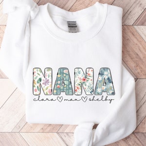 以下が含まれることがあります： 白いスウェットシャツに、花柄の文字で「NANA」と書かれています。「NANA」の下には、「Clara」、「Max」、「Shelby」という名前が、ハートマークで区切られて書かれています。