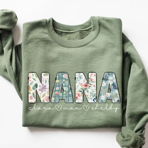 以下が含まれることがあります： 花柄の文字で「NANA」と書かれた緑色のスウェットシャツ。 「NANA」の下には、「Clara」、「Max」、「Shelby」という名前が筆記体で書かれています。