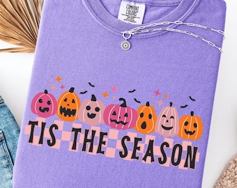 May include: A violet Comfort Colors sweatshirt featuring a graphic of smiling pumpkins and the phrase "TIS THE SEASON" in black. The design includes bats and stars. A silver necklace and jeans are also visible.
