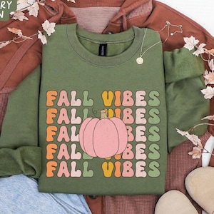 May include: A military green sweatshirt with the text "FALL VIBES" in a retro font and a pink pumpkin graphic. The sweatshirt is displayed with a brown hoodie, blue jeans, and tan shoes, suggesting a fall theme.