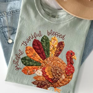 May include: A light green t-shirt with a colorful turkey design and the words "grateful, thankful, blessed." The turkey has a patchwork design with red, orange, and brown feathers. The shirt is paired with blue jeans and a tan hat.