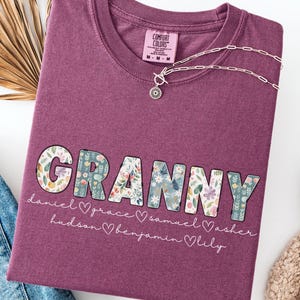 May include: A maroon Comfort Colors t-shirt with the word "GRANNY" in a floral pattern. Below the word are names and hearts. A silver necklace with a pendant is on the shirt. The shirt is next to a pair of blue jeans and a white shoe.