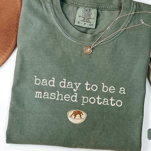 May include: Moss green Comfort Colors t-shirt with the phrase "bad day to be a mashed potato" in white script. A gold necklace with a small pendant and a brown hat are also visible.